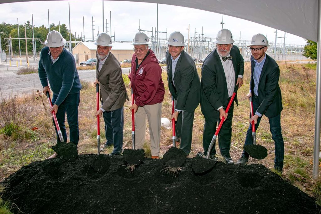 Group of men each with a shovel digging up dirt in front of a storage site.