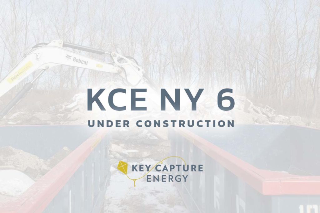 The words KCE NY 6 under construction with Key Capture Energy logo underneath it, construction site behind.