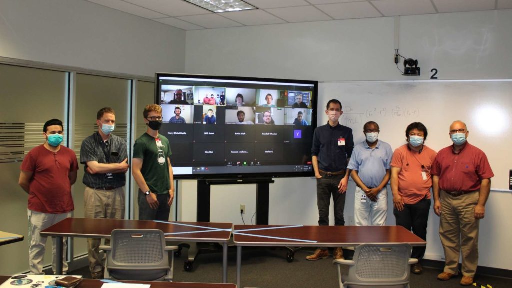Group of employees standing by a large screen with a video call open.