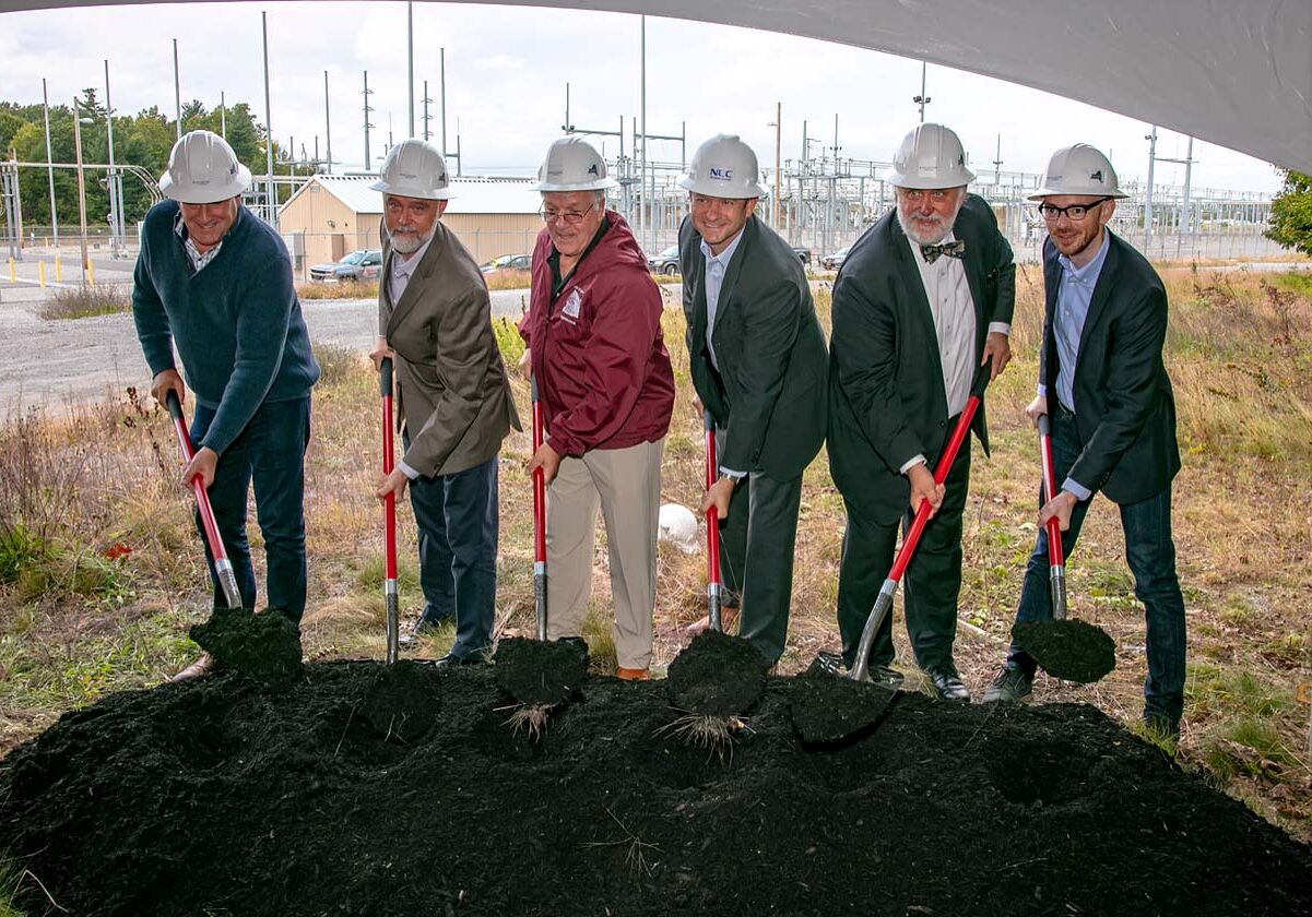 Group of men each with a shovel digging up dirt in front of a storage site.