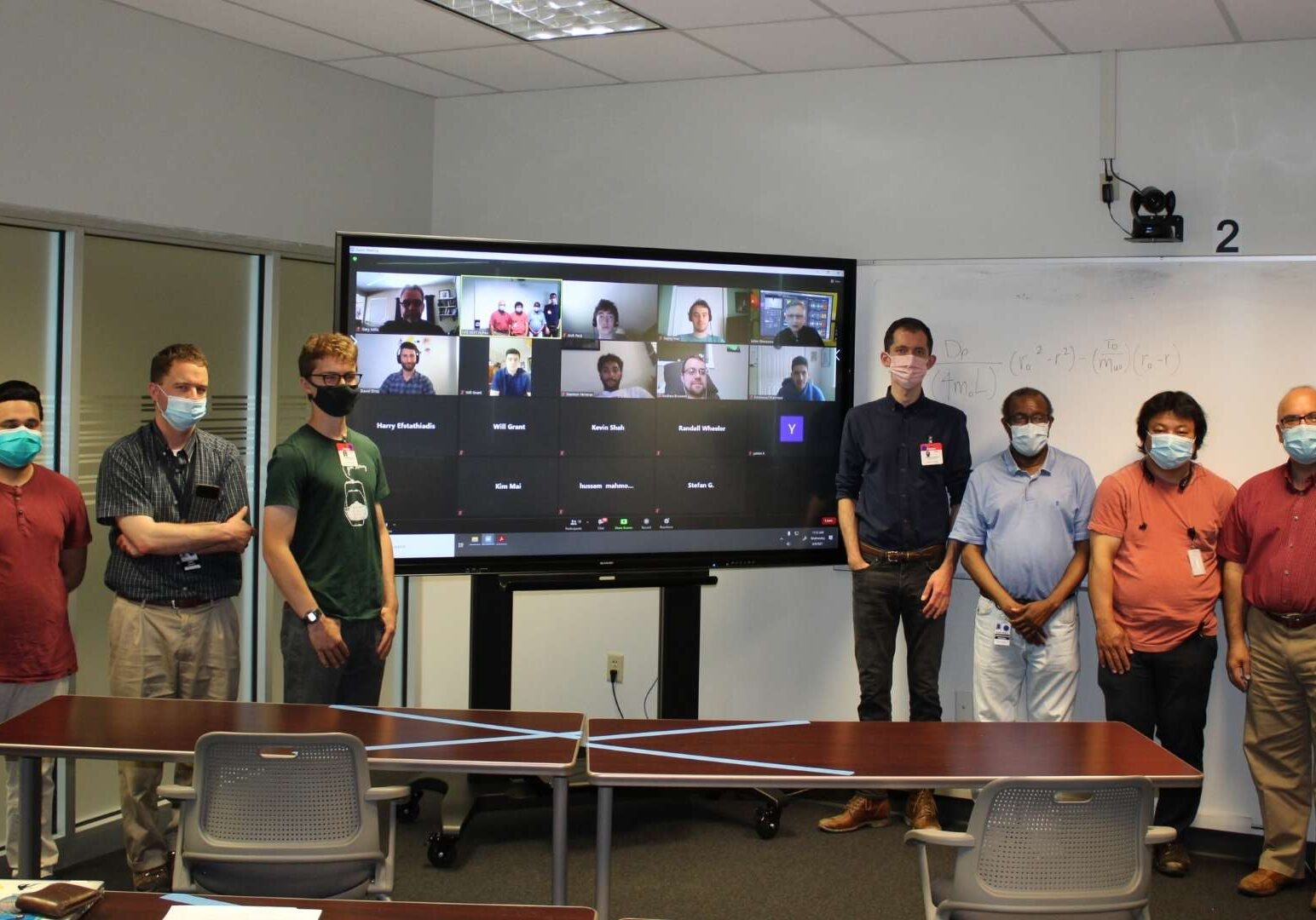 Group of employees standing by a large screen with a video call open.